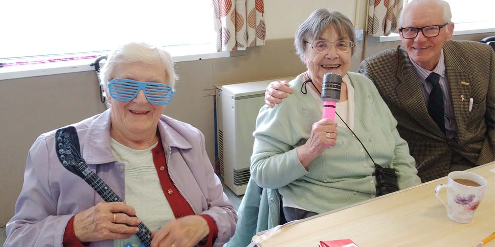 3 elderly people smiling on chairs holding inflatable microphones and wearing novelty glasses